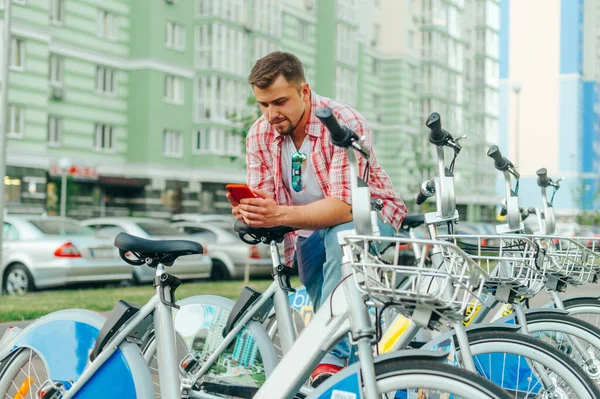 Concentrated man in casual clothes stands near a row of bicycles and shares in a program on a smartphone. Guy with a beard stands by a rented bicycle and uses a smartphone