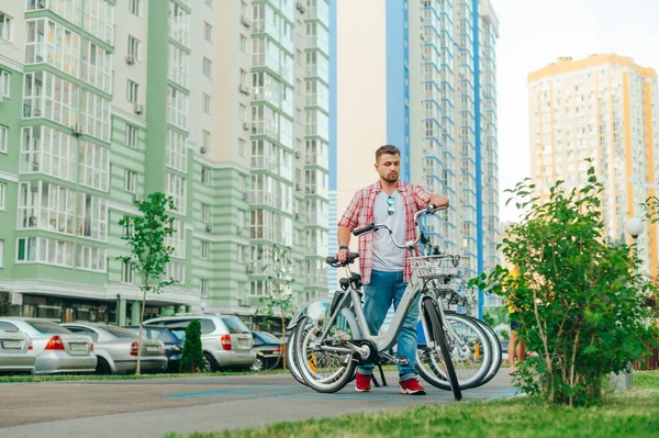 Man in casual clothes with rented city bike on cityscape background. A man with a beard rented a bicycle to walk around the city. Tourist pushes a bicycle out of the sharing