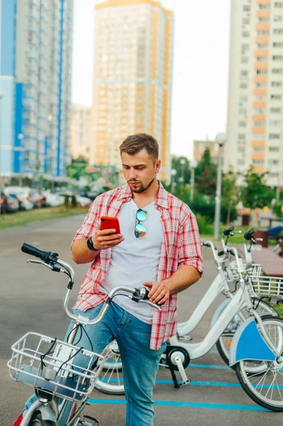Sakallı ve tişörtlü yetişkin bir adam elinde akıllı bir telefonla arka plandaki bisiklet sırasına karşı duruyor ve bisiklet paylaşmak için mobil bir program kullanıyor. Şehir bisikleti sürmek