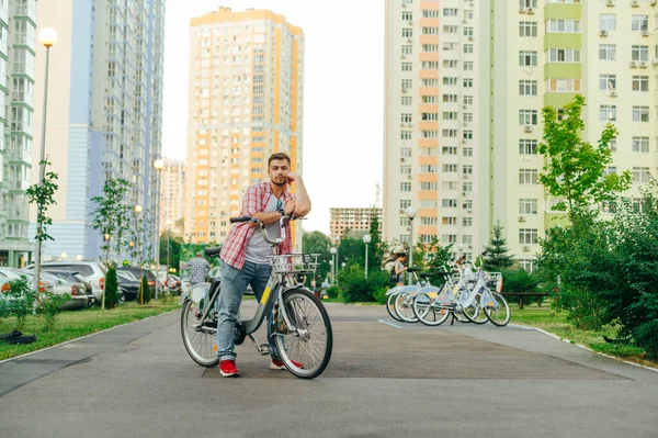 Portrait of a man with a city bike on the background of a row of bicycles for sharing and cityscape, standing on the path and looking at the camera with a serious face. Evening bike ride