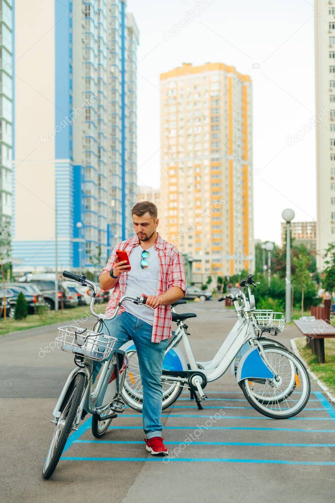 Adult man in a shirt and with a beard sits on a shering bicycle and uses a smartphone. Tourist rents a bicycle through a mobile application. Eco transport. Vertical