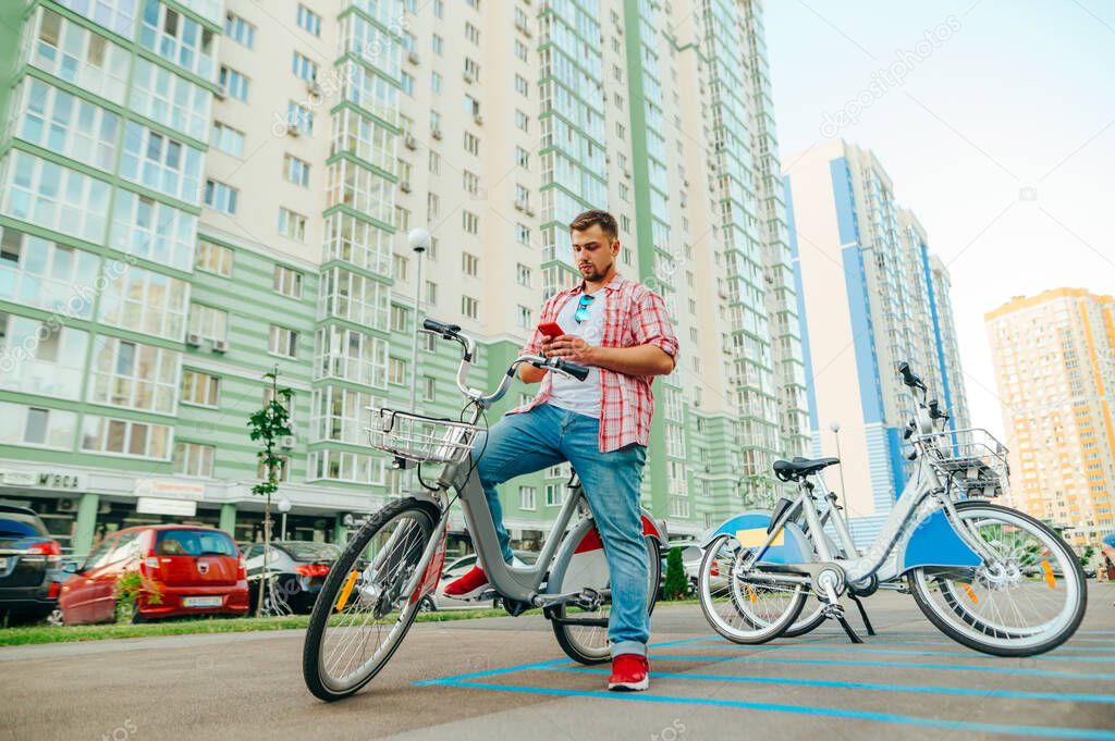 Adult man in a shirt and with a beard sits on a shering bicycle and uses a smartphone. The tourist rents a bicycle through a mobile application. Eco transport