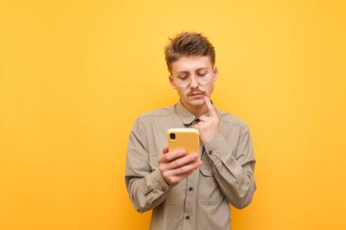 Bıyıklı ve gözlüklü düşünceli genç adam sarı arka plana karşı akıllı telefon kullanıyor, ekrana bakıyor ve tişört giydiğini düşünüyor. Ciddi yüzlü komik inek telefonu kullanıyor. İzole edilmiş