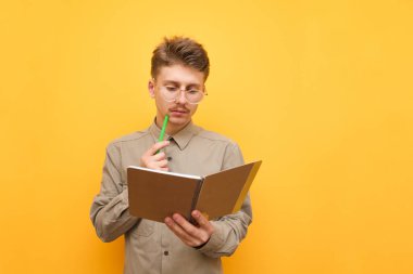 Elinde not defteri ve kalemle ciddi bir öğrencinin portresi, dikkatle deftere bakıyor ve görevi düşünüyor. Sarı arka planda izole edilmiş kalem ve not defteri olan tişörtlü ve gözlüklü inek. Boşluğu kopyala