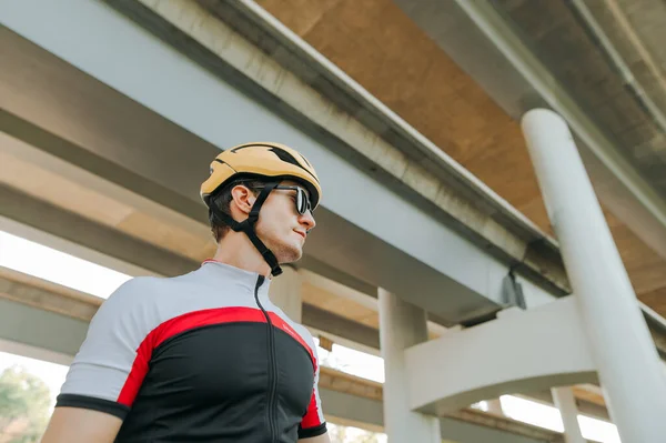 Close up portrait of professional cyclist in sports outfit stands on the background of the bridge and looks to the side, bottom view.Portrait of serious handsome man cyclist on architecture background