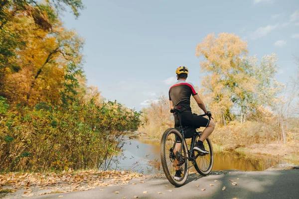 Professional cyclist in helmet and sportswear stands with bicycle, background of lake and autumn park and admiring the landscape, view from the back.Cyclist looking at a beautiful autumn landscape