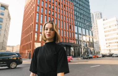 Şık siyah elbiseli çekici bayan modern bir metropolün arka planında duruyor, ciddi bir yüzle kameraya bakıyor. Siyah gömlekli şık kız kameraya poz veriyor.