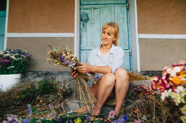 Yetişkin bir kadın ülkede bir buket güzel kurumuş çiçek üretiyor. Bir kır evinin duvarının arkasındaki sarışın çiçekçi büyümüş kurumuş çiçeklerden buketler yapıyor..