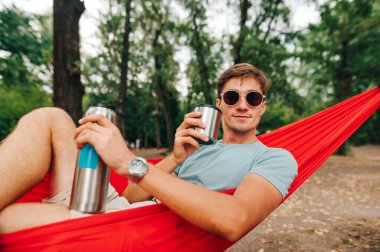 Pozitif genç adam ormanın ortasında bir hamakta yatıyor, termostan alkollü bir içecek içiyor ve yüzünde bir gülümsemeyle kameraya bakıyor..