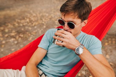 Güneş gözlüklü genç adam parkta bir hamakta uzanıyor ve bir fincandan su içiyor. Ormanda bir hamakta dinlenmek.