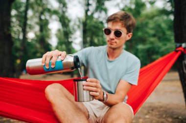 Günlük giysiler ve güneş gözlüklü genç adam parkta bir hamakta oturuyor ve gümüş bir termostan bir kaba soğuk su döküyor..