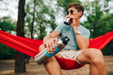 Parlak elbiseli genç adam parkta bir hamakta oturuyor ve gümüş bir termostan su içiyor. Termosa odaklan..