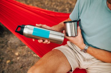 Adam kırmızı bir hamakta oturuyor ve elinde gümüş bir termos tutuyor. Termos bir erkek turistin elinde, bir hamakta dinleniyor..