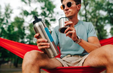 Genç adam elinde bir termos soğuk içecekle ormanda bir hamakta oturuyor. Turist doğada bir hamakta dinleniyor ve çay içiyor..