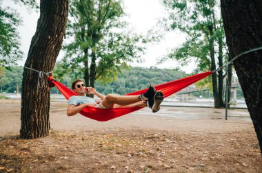 Parktaki bir hamakta dinlenen mutlu bir adam ve şehrin arka planına karşı telefonla arıyor gezinti güvertesi ve köprüyle.