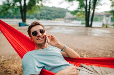Güneş gözlüklü yakışıklı bir adam ağaçtaki bir hamakta uzanıyor ve telefonda yüzünde bir gülümsemeyle konuşuyor. Ormanda bir hamakta uzanmak zevklidir..