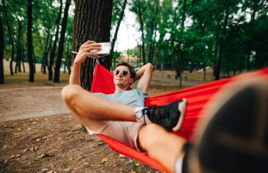 Hamakta yatarken, günlük kıyafetleriyle selfie çeken genç bir adam. Ormandaki bir ağaca bağlanmış hamakta dinlenen ve akıllı telefon kullanan bir adam..