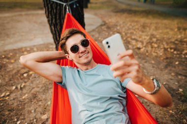 Güneş gözlüklü mutlu adam parkta bir hamakta uzanıyor ve yüzünde bir gülümsemeyle akıllı bir telefondan selfie çekiyor. Turist elinde akıllı bir telefonla bir hamakta dinleniyor..