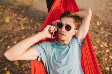 Güneş gözlüklü ciddi bir gencin portresi ve parktaki bir hamakta uzanmış, odaklanmış bir yüzle konuşan gündelik giysiler..