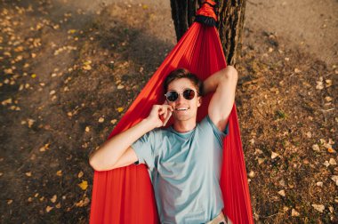 Sun Colari 'deki mutlu genç bir adamın portresi ve tişört parkta kırmızı bir hamakta yatıyor, kameraya bakıyor ve telefonla arıyor..