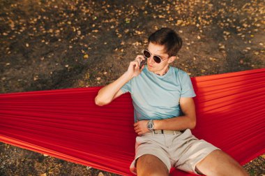 Güneş gözlüklü ciddi bir gencin portresi ve parktaki bir hamakta uzanan ve odaklanmış bir yüzle konuşan gündelik giysiler..