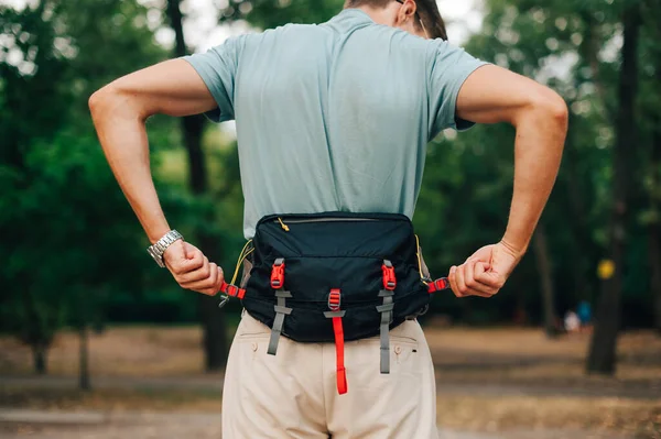 Man in casual clothes stands in the woods during the day and fastens fastex on a waist bag. Tourist with a waist bag on his belt stands in the woods. Back view.