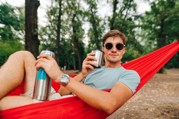 Pozitif genç adam ormanın ortasında bir hamakta yatıyor, termostan alkollü bir içecek içiyor ve yüzünde bir gülümsemeyle kameraya bakıyor..