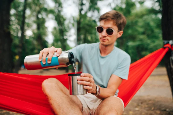 Günlük giysiler ve güneş gözlüklü genç adam parkta bir hamakta oturuyor ve gümüş bir termostan bir kaba soğuk su döküyor..
