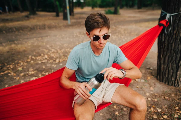 Güneş gözlüklü yakışıklı ormanda kırmızı bir hamakta oturuyor ve termosun kapağını açıyor, sıcak çay içmeye gidiyor..