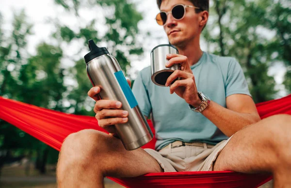 Genç adam elinde bir termos soğuk içecekle ormanda bir hamakta oturuyor. Turist doğada bir hamakta dinleniyor ve çay içiyor..