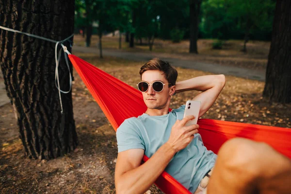 Güneş gözlüklü ve günlük kıyafetli yakışıklı bir adam ormanda bir hamakta elinde akıllı bir telefonla uzanıyor ve konsantre bir yüzle başka yere bakıyor. Parkta dinlen..