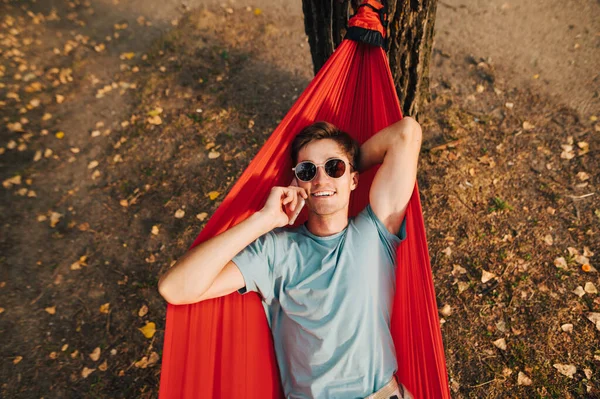 Sun Colari 'deki mutlu genç bir adamın portresi ve tişört parkta kırmızı bir hamakta yatıyor, kameraya bakıyor ve telefonla arıyor..