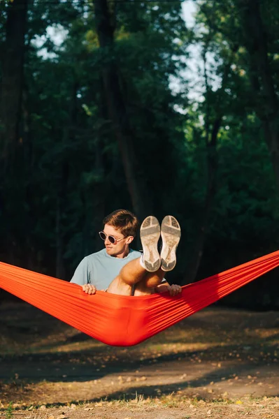 Güneş gözlüklü genç adam ve günbatımında ormanda kırmızı bir hamakta sallanan gündelik giysiler. Dikey.