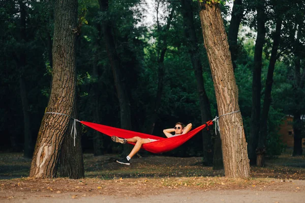 Genç adam gün batımında parkta kırmızı bir hamakta yatıyor. Adam ormanın arka planında bir hamakta dinleniyor..