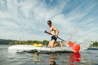 Atletik genç adam nehir kenarındaki bir panoda aktif olarak kürek çekiyor ve kameraya poz veriyor. Nehirdeki bir kürek tahtasında antrenman..