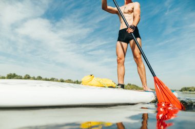 Geçmişi. Kesilmiş fotoğrafı kapat. Kaslı vücutlu atletik genç bir adam yemek tahtası ve küreklerin üzerinde duruyor. Güneşli bir günde suda aktif eğlence..