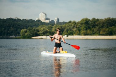 Kaslı vücutlu atletik bir adam güzel bir manzaranın arka planında nehrin üzerinde süzülüyor. Güneşli bir yaz gününde bir kürek tahtasında antrenman yapmak.