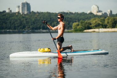 Güneş gözlüklü atletik yakışıklı adam ve güzel bir manzaranın arka planında nehirde yemek masasında kürek çeken adam. Gölde bir sörf tahtasıyla aktif eğlence..