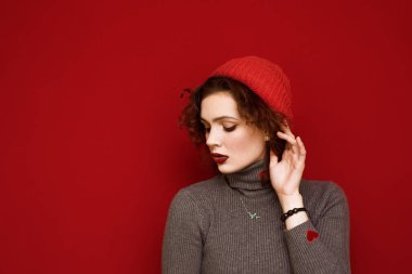 Şapkalı ve kıvırcık saçlı çekici genç manken kameraya ciddi bir yüzle bakıyor, kırmızı bir arka plana bakıyor. Kırmızı arka planda şık bir kızın moda portresini kapat.