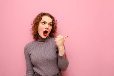 Şok olmuş, mutlu yüzlü bir kız pembe arka planda duruyor, bakışlar ve noktalar fotokopi alanında. Kıvırcık, şaşırmış bir kız baş parmağını tek başına gösteriyor..