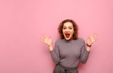 Gri balıkçı yaka kazaklı, şok olmuş kıvırcık kız portresi kameraya bakıyor ve mutlulukla bağırıyor. Neşeli bayan pembe arka planda kazanmaktan duygusal olarak memnun. Boşluğu kopyala