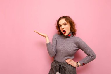 Kıvırcık saçlı kadın pembe arka planda duruyor ve elini fotokopi alanına doğrultuyor, kameraya şok olmuş gibi bakıyor. Duygusal kız reklam için bir yer tutuyor..