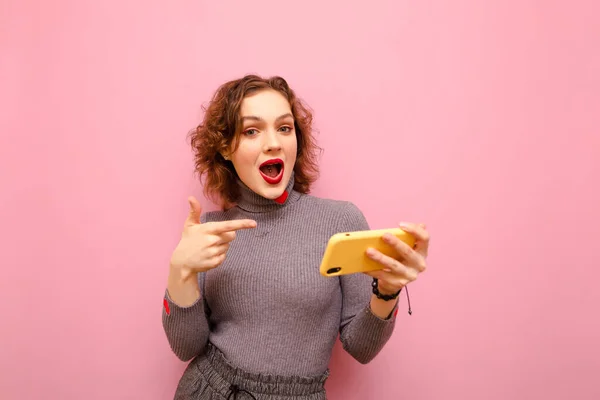 Happy girl in pink clothes stands with a smartphone in her hand on a pink background and shows a finger on the gadget with a smile on face.Joyful lady with surprised face shows finger on phone in hand