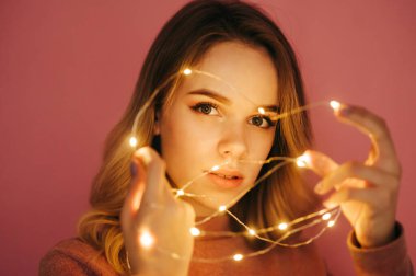 Süveter giymiş güzel bir bayan pembe arka planda duruyor ve ellerinde Noel ışıkları var. Pembe arka planda çelenkle oynayan bir kadın..