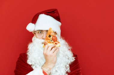 Noel Baba 'nın gözünün önünde bir dilim pizza tutarken yakın plan portresi ve kırmızı arka planda izole edilmiş ciddi bir yüzle kameraya bakarken. Boşluğu kopyala