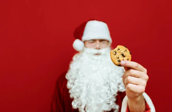 Photo of Santa Claus on a red background holding a cracked cookie with chocolate in his hand close up. Christmas background with cookies and Santa, isolated on red. New Year concept.