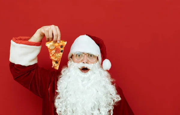 Shocked Santa Claus with a slice of pizza in his hand isolated on red background, surprised looking into the camera, empty space to the right. Portrait of surprised santa with piece of delicious pizza