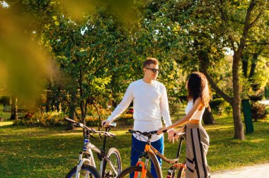 Güzel genç kadın ve erkek, sonbahar parkında durup ellerinde bisikletlerle iletişim kuruyorlar. Çift parkta bisiklet sürüyor..