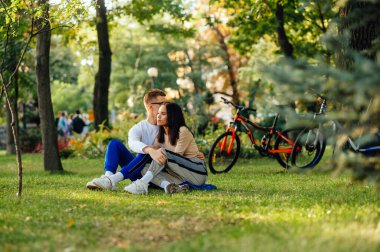 Güzel genç bir çift haftasonunu parkta çimlerin üzerinde oturup bisiklet arka planında kucaklaşarak geçirir. Erkek ve kadın bisiklet sürdükten sonra sonbahar parkında çimlerde dinleniyorlar..