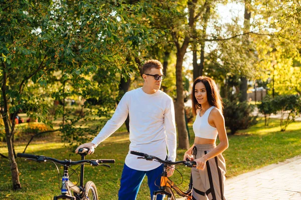 秋の公園を自転車で歩き 話をする明るいカジュアルな服を着た美しい女性と男性 公園で自転車を遊んでいる若いカップル — ストック写真